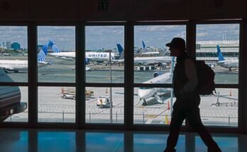 Un viaggiatore che indossa uno zaino e un cappello viaggia all'aeroporto internazionale di Newark Liberty all'inizio di giugno. Attraverso le grandi finestre dietro di lui, puoi vedere diverse United Airlines parcheggiate a Gates.